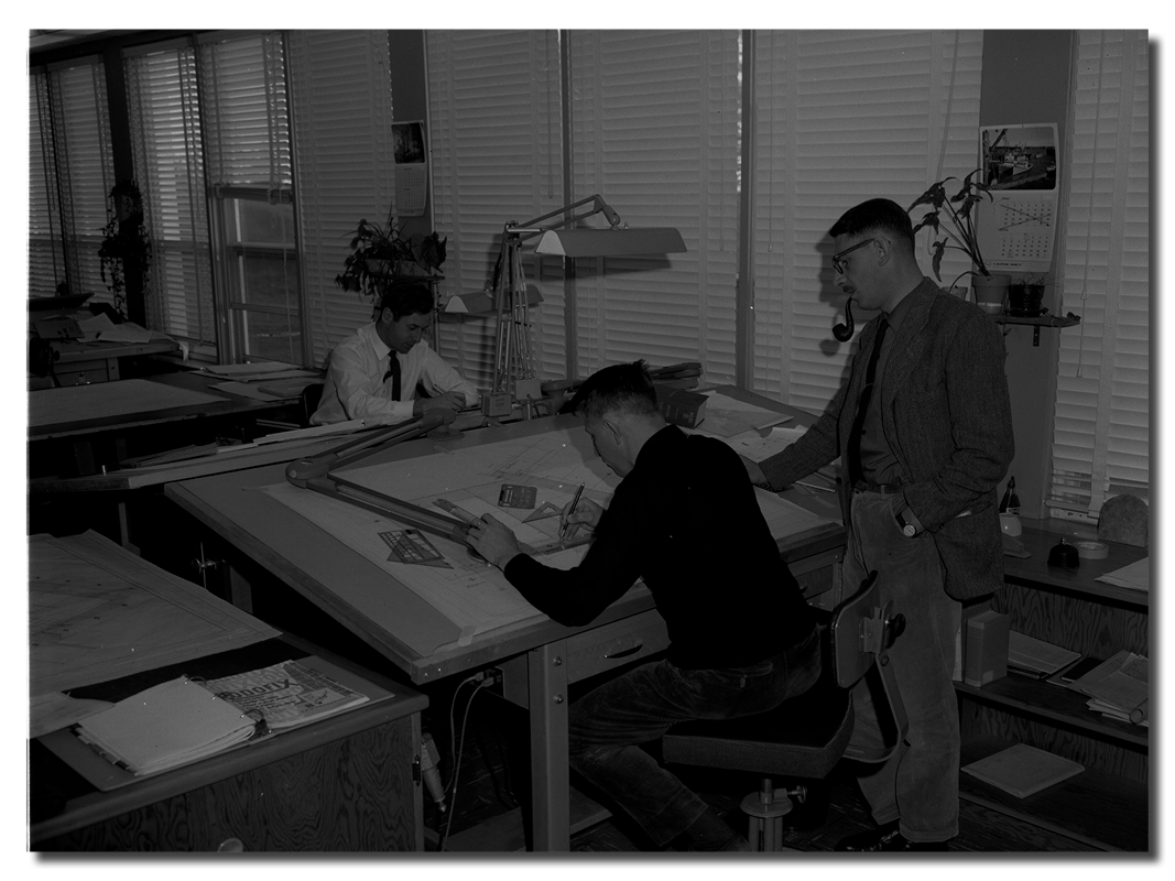 Black and white image of three people bent over a drafting table.